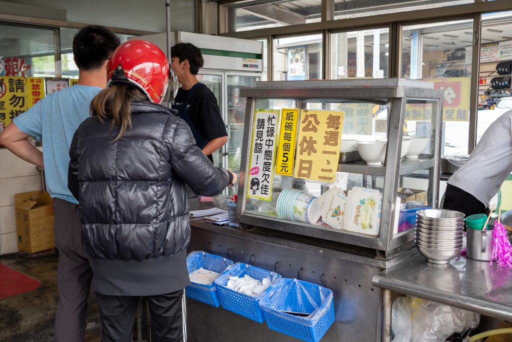 客人在王家牛肉湯的櫃檯前排隊點餐,告示寫著每週一二公休、紙碗每個5元