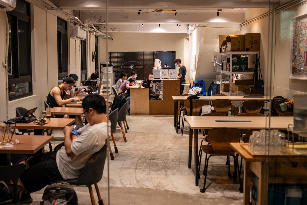 二樓室內全景，中間是原木大長桌與兩側座位，客人各自在筆電前工作，最裡面是吧檯