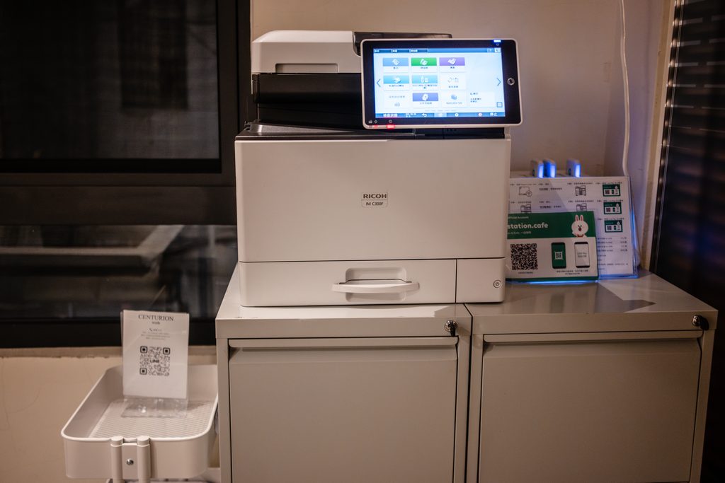 白色 RICOH 影印機，機身上放著一台顯示操作介面的平板，旁邊櫃子上有說明小卡