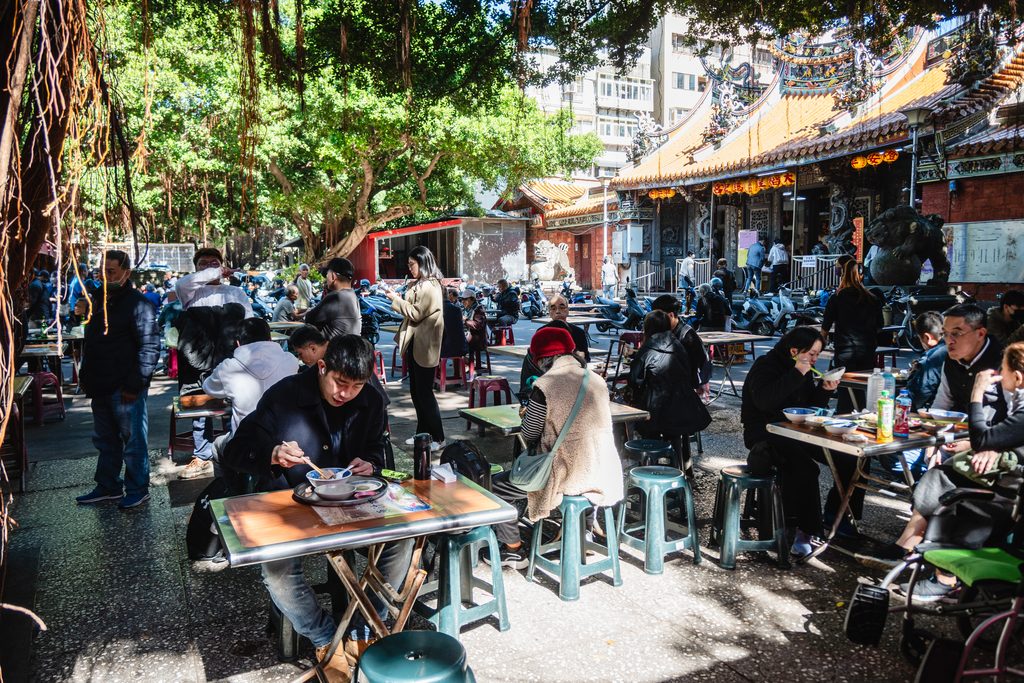 大稻埕慈聖宮廟埕廣場，老榕樹下擺滿桌椅，民眾坐著用餐，後方是慈聖宮廟頂