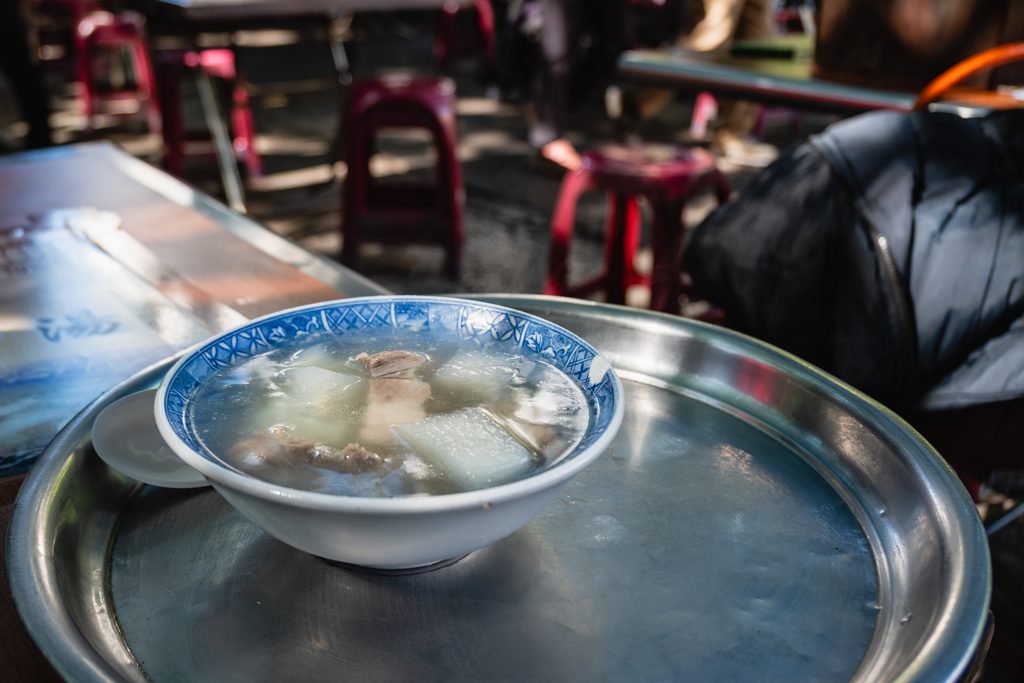 不鏽鋼托盤上的一碗原汁排骨湯，青花瓷碗盛裝，背景是廟埕的桌椅