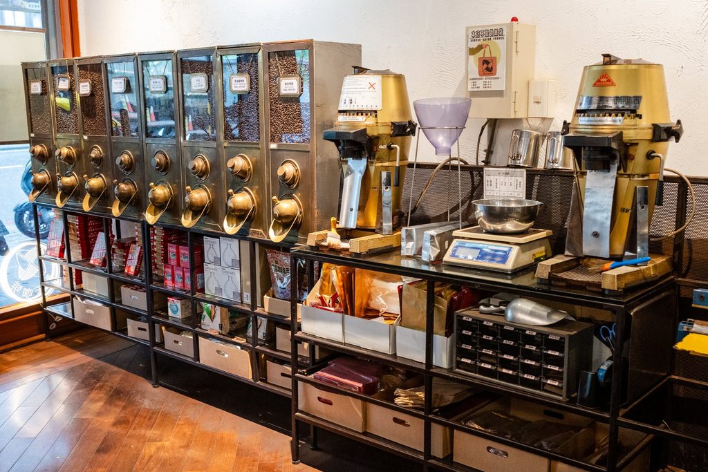 店內一側的烘焙與販售區,左邊是整排銅色出豆口的豆倉,中間兩台黃色的營業用磨豆機與電子秤,下層層架擺著掛耳包與商品抽屜