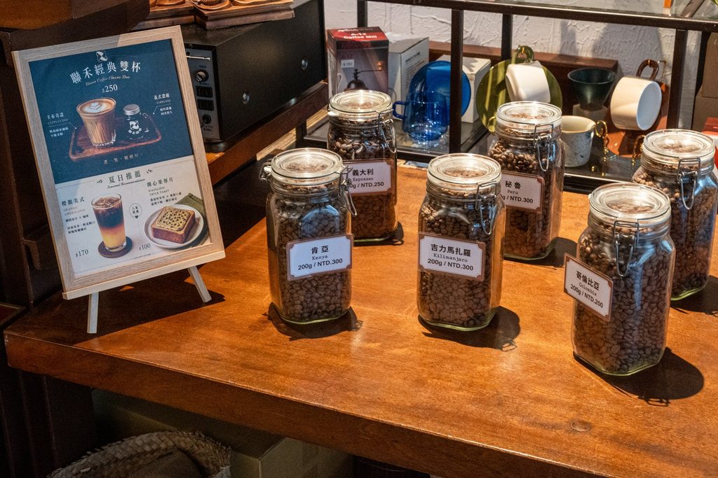 木桌上一排密封玻璃罐裝著烘好的咖啡豆,每罐前面立著品名與 200 公克價格的小牌,桌邊立著一張店內的飲品宣傳卡